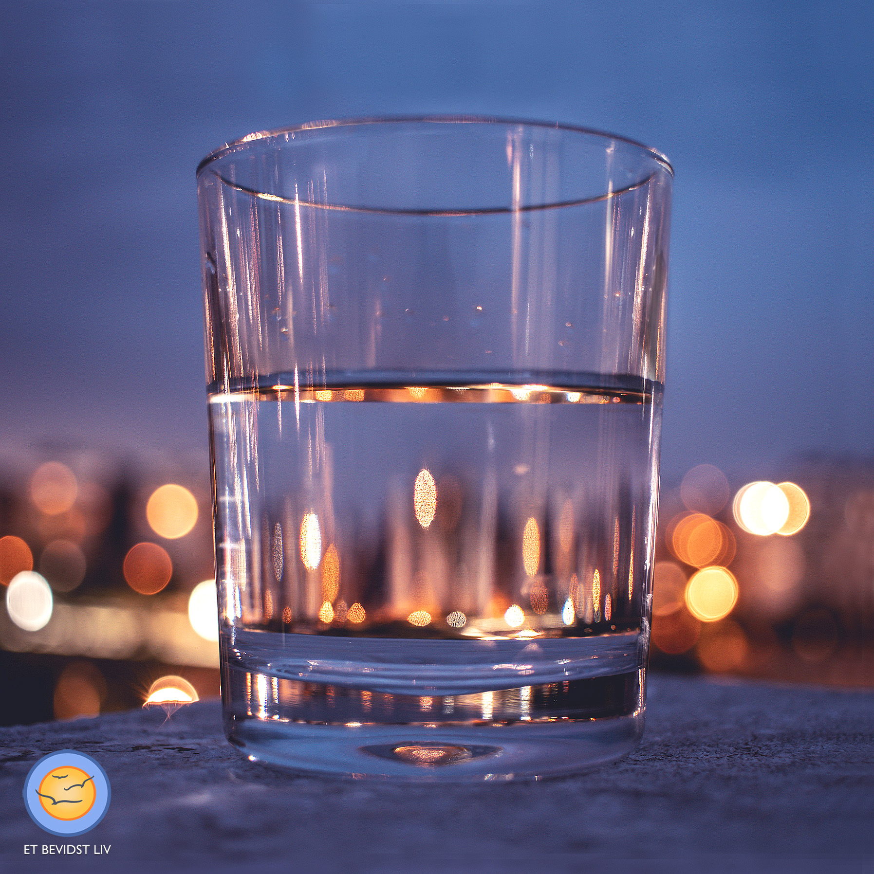 Foto af et halvt fyldt (eller halvt tomt) glas ved nattetide, med refleksioner af byens lys.
